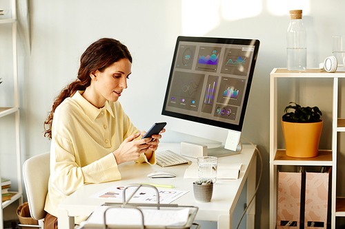 Stylish young adult Caucasian woman with long hair wearing casual beige shirt sitting in modern office scrolling news feed on smartphone