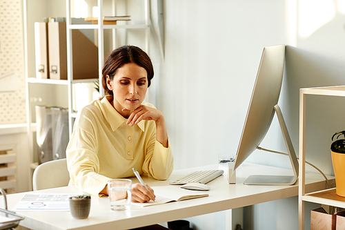 Elegant young adult Caucasian entrepreneur in oversized shirt sitting at table in modern office writing notes in notebook