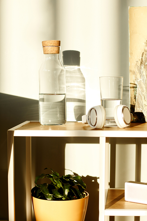 Vertical minimalistic still life composition of bottle, headphones and glass of water on table in daylight, modern office interior