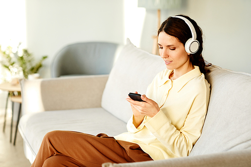 Young adult Caucasian woman wearing headphones sitting on couch at home choosing music to listen to on smartphone