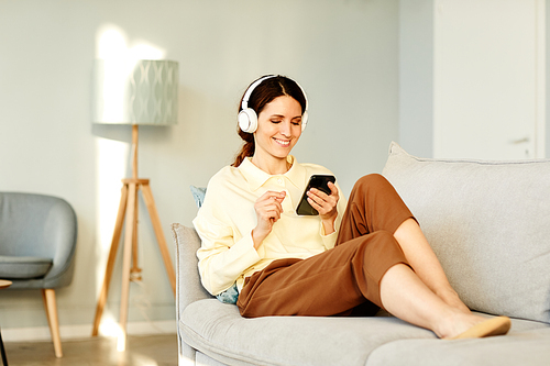 Attractive young adult Caucasian woman wearing casual clothes relaxing on couch at home listening to music in headphones