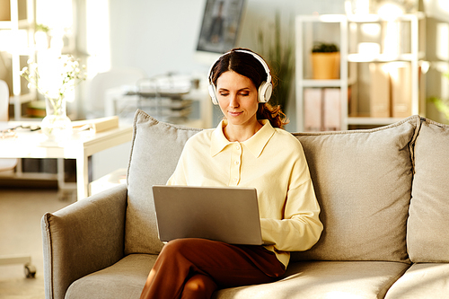 Moden young adult Caucasian woman sitting on couch in living room listening to music in wireless headphones and searching something in Internet on laptop