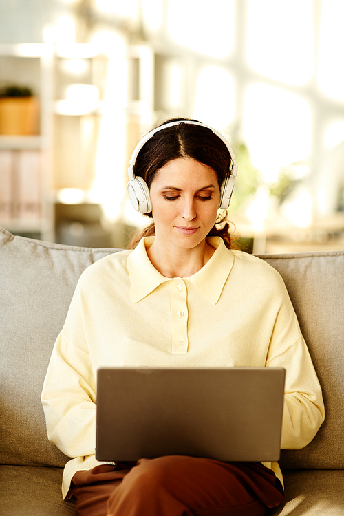 Vertical shot of beautiful young adult Caucasian woman wearing casual clothes and headphones sitting on sofa at home working remotely using laptop