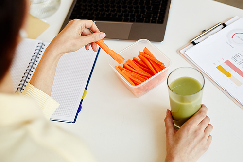 High angle view of unrecognizable woman working in office having fresh carrot sticks and vegetable green smoothie for snack