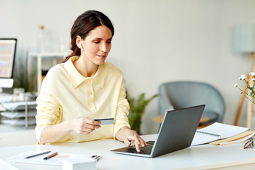 Horizontal medium shot of young adult Caucasian woman sitting at desk in office using laptop and bank card to order food delivery