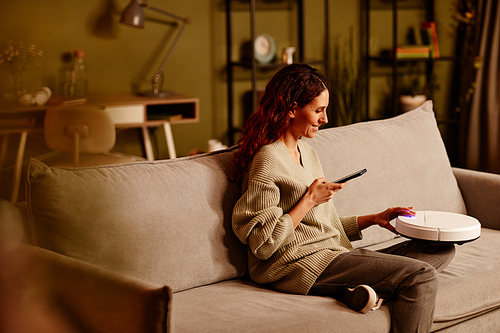 Cheerful young adult Caucasian woman holding smartphone adjusting settings of modern robotic vacuum cleaner