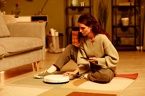 Young adult Caucasian woman wearing casual clothes sitting on floor in living room turning on robotic vacuum cleaner