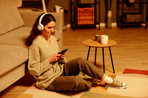 Young adult Caucasian woman wearing causal outfit and wireless headphones sitting relaxed on floor in living room at home choosing playlist