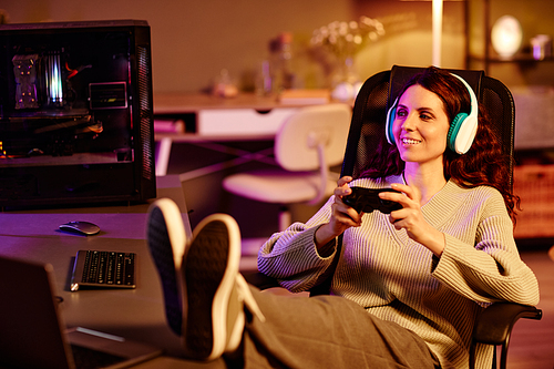 Modern young adult Caucasian woman wearing casual clothes and headphones spending evening playing video games with use of controller