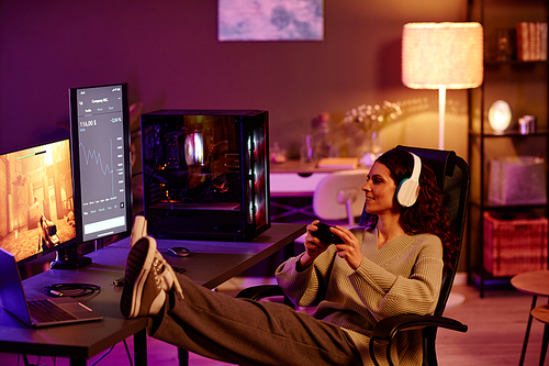 Modern Caucasian woman wearing casual clothes and headphones sitting with feet on desk spending evening playing video games with use of controller