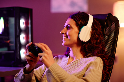 Modern young adult woman with long curly hair wearing headphones spending evening at home enjoying playing video games