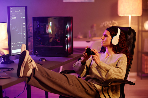 Young adult woman wearing comfortable clothes sitting at desk with modern computers on it playing video games with console at night