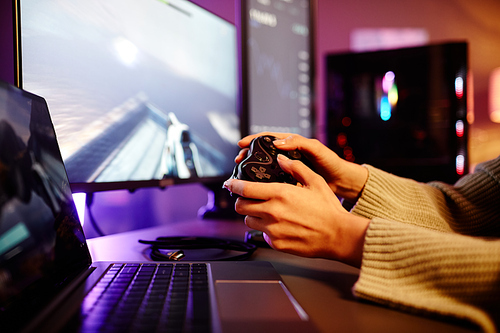 Unrecognizable woman sitting at desk with computer and laptop on it playing shooter video game at night