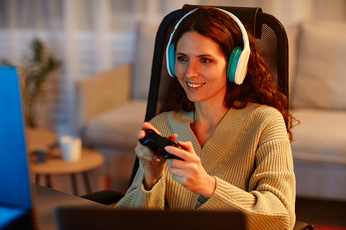 Cheerful young adult Caucasian woman wearing headphones playing video game using controller in living room late in evening