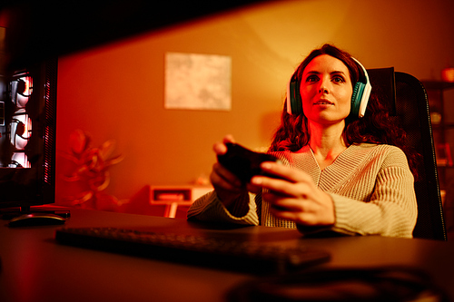 Young adult Caucasian woman with long curly hair wearing headphones enjoying playing video game at night