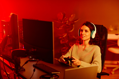 Joyful young adult Caucasian woman playing shooter or racing video game using console controller in living room at night, neon red light