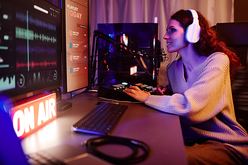 Young adult Caucasian woman with long curly hair wearing casual outfit and headphones recording podcast or broadcast in evening