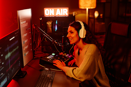 Joyful young adult Caucasian woman with long curly hair wearing casual outfit and headphones telling stories to listeners during radio broadcasting at night