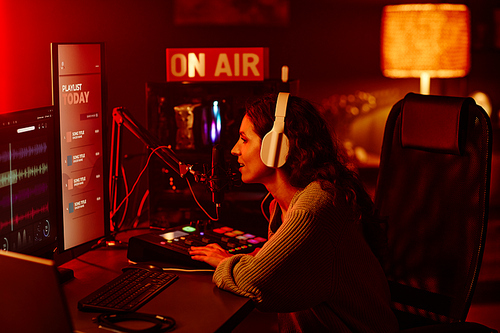 Modern Caucasian woman with long curly hair wearing casual outfit and headphones recording podcast or broadcast at night