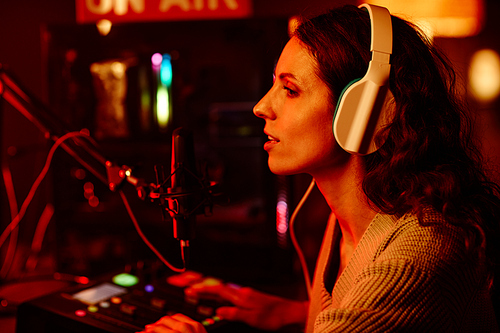 Side view of attractive young adult Caucasian woman Young adult with long curly hair wearing headphones telling stories while recording podcast or broadcast at night