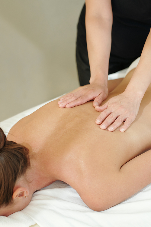 Hands of massage therapist touching back of young woman lying on white sheets in luxurious spa salon