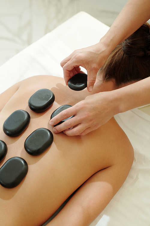 Hands of masseuse putting hot spa stones on female back during healthcare and beauty procedure in luxurious salon