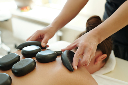 Part of body of young female with hot spa stones on her back and professional masseuse holding two of them