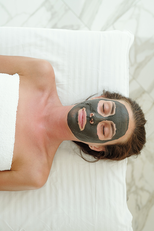 Overview of young woman with clay mask on her face lying on white sheet in luxury beauty salon at spa resort