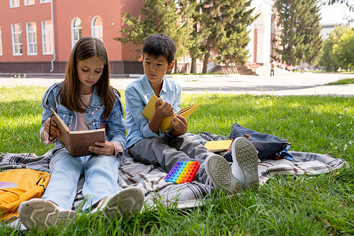 Two cute intercultural schoolchildren reading books while resting on green lawn