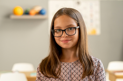 Cute secondary school pupil in eyeglasses looking at camera while standing in classroom