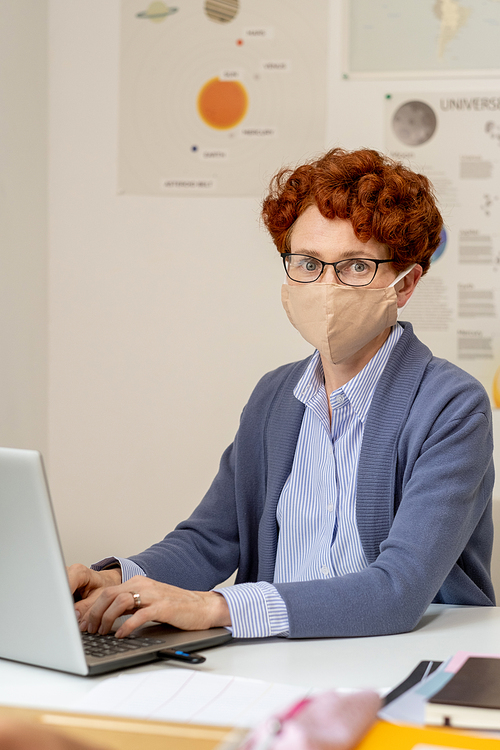 Contemporary school teacher in protective mask and eyeglasses networking in front of laptop