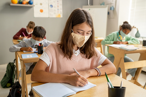 Cute schoolgirl and her classmates in protective masks making notes in their copybooks