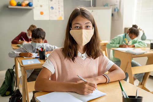 Adorable schoolgirl in protective mask looking at you while making notes at lesson