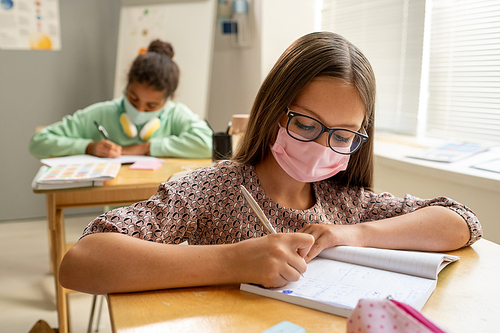 Cute diligent schoolgirl in protective mask making notes in copybook at lesson
