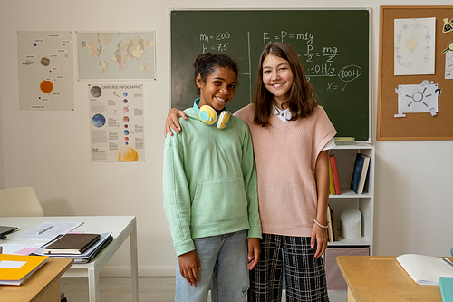 Two cheerful multiracial schoolgirls in casualwear standing against blackboard