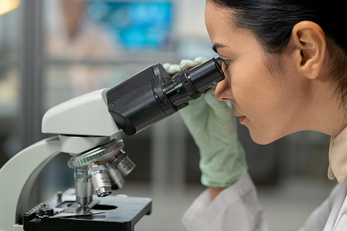 Young female scientist in protective gloves looking in microscope while studying new chemical substance and its characteristics