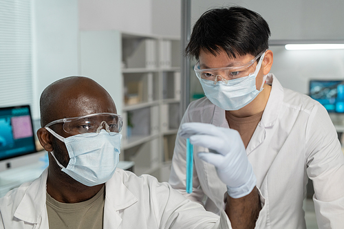 Two young intercultural scientists in lab coats, gloves and protective masks looking at sample of blue liquid in flask