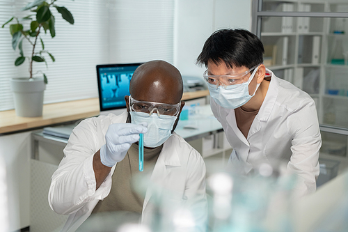 Two young interracial chemists in lab coats and protective masks looking at blue liquid in test tube while studying new compound