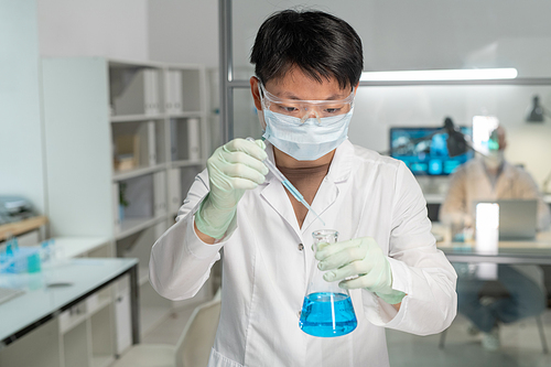 Young Asian female chemist adding drop of chemical substance into blue liquid while carrying out scientific experiment in laboratory