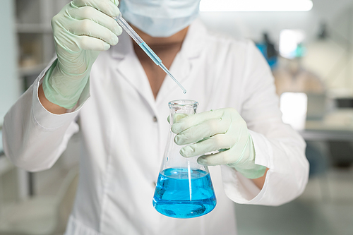 Contemporary scientist in surgical gloves, mask and lab coat adding drop of liquid into flask with blue chemical substance