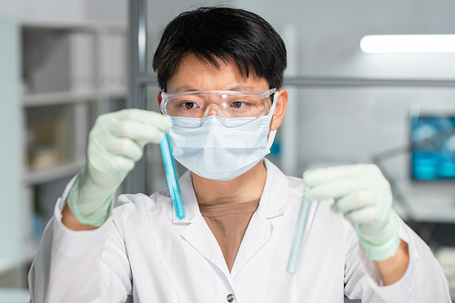 Asian female scientist in protective eyeglasses, mask and gloves comparing two samples of liquid substances