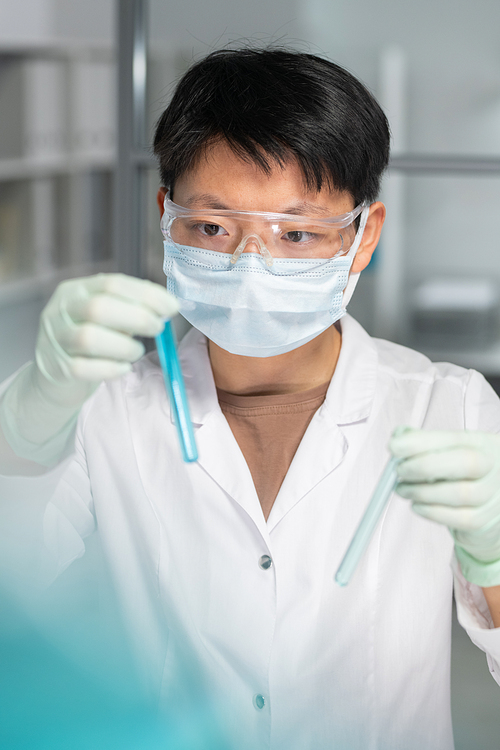 Young female scientist of Asian ethnicity holding flask with blue liquid in hand and looking at it during scientific research