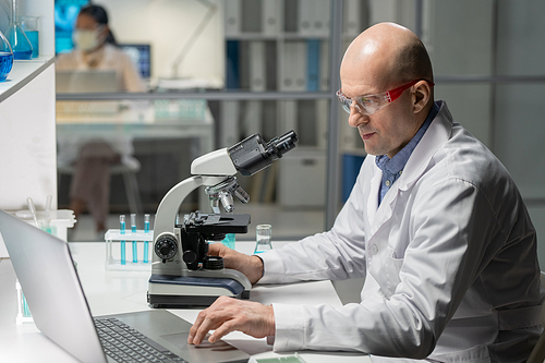 Bald mature chemist in lab coat entering data in computer while using microscope during scientific experiment or research