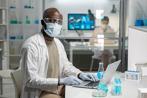 Young African American male scientist in protective gloves, mask and eyeglasses typing on lapto pkeyboard during work in laboratory