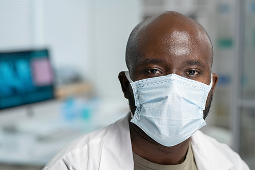 Head of young black man in whitecoat protective mask looking at camera while sitting by workplace in modern scientific laboratory