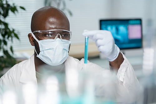 Young black man in lab coat, protective mask and gloves looking at flask containing blue liquid during work in laboratory