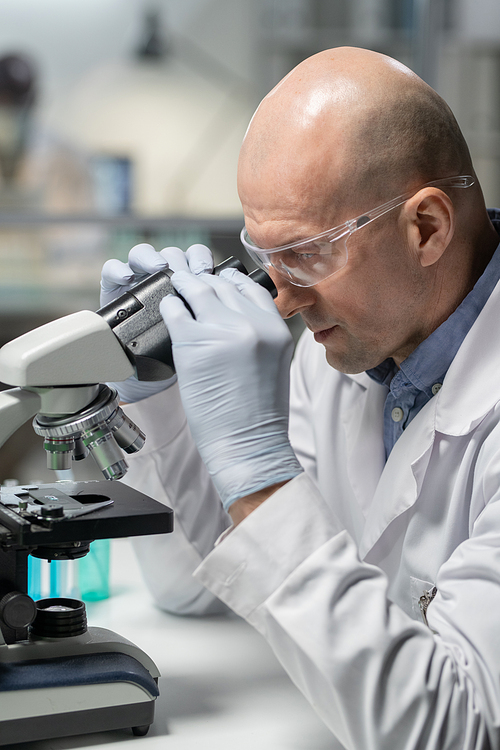Side view of bald mature man in whitecoat, gloves and eyeglasses using microscope while studing new chemical substance in laboratory