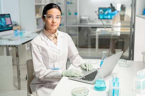 Young contemporary female chemist with laptop entering new scientific data while sitting by workplace and looking at camera
