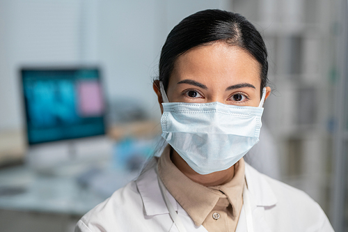 Young pretty female clinician in whitecoat and protective mask on face looking at camera while standing in medical office