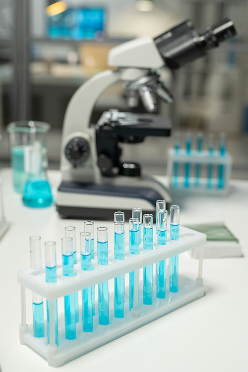 Group of flasks with samples of blue liquid standing on workplace of scientist against microscope and other glass equipment